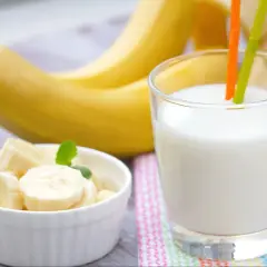 Szklanka pysznego mleka bananowego z dwoma słomkami, obok miseczka z pokrojonymi bananami i całe banany w tle.
