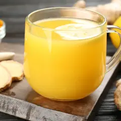 Napój z cytryny i imbiru, który może pomóc na skurcze nóg. Obok szklanki z żółtym płynem leżą plastry imbiru, cytryna i miseczka z kurkumą.