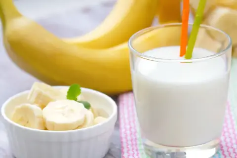 Szklanka pysznego mleka bananowego z dwoma słomkami, obok miseczka z pokrojonymi bananami i całe banany w tle.