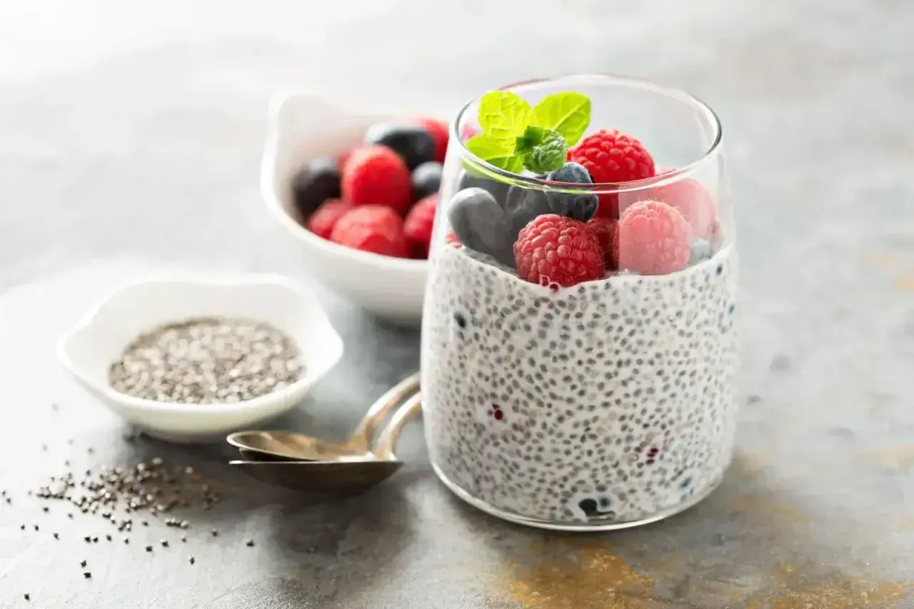 Pudding chia z owocami leśnymi, pokazujący jak przygotować nasiona chia. Obok miseczka z nasionami i łyżeczki.