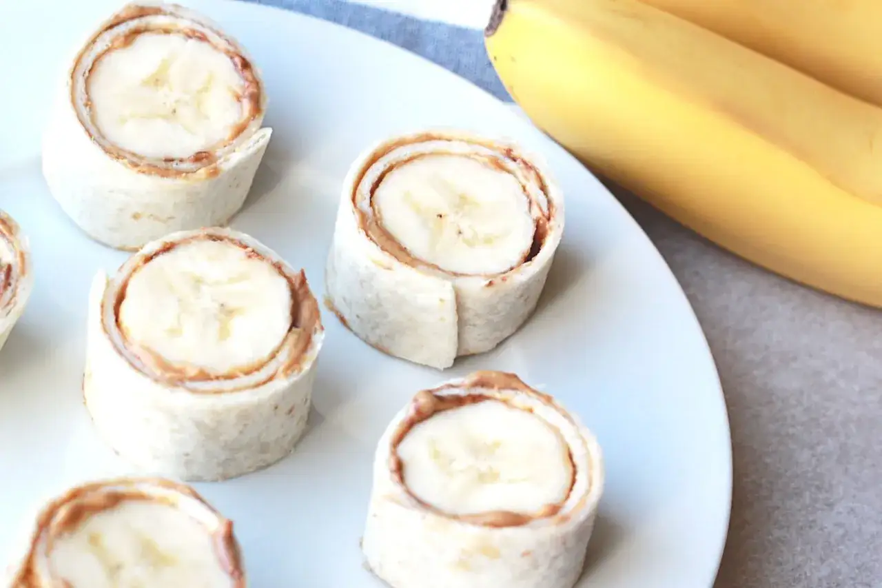 Roladki z tortilli z bananem i masłem orzechowym, idealne jako banan na noc. Obok leżą świeże banany.