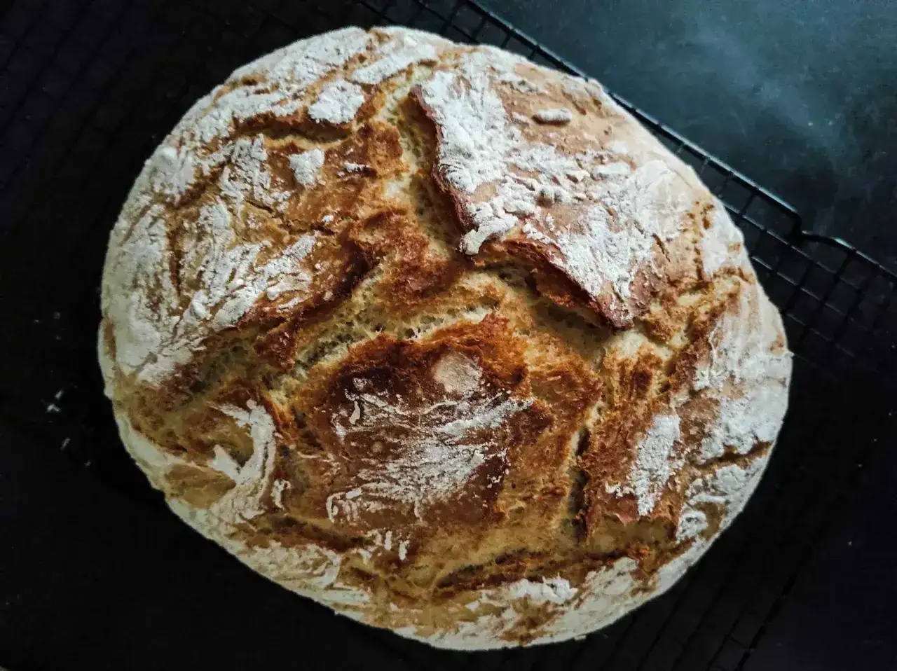 Chleb rustykalny z chrupiącą skórką i puszystym wnętrzem, posypany mąką, leży na czarnej kratce.